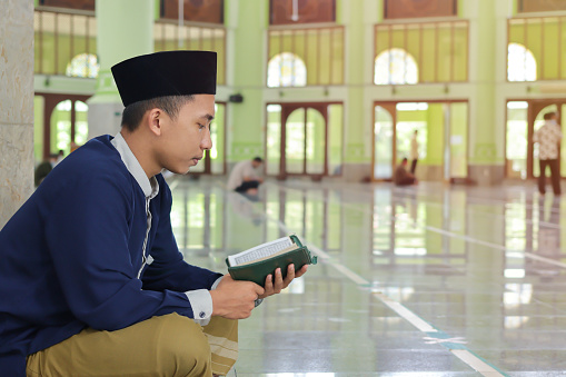 Religious Asian man in muslim shirt and black cap reading the holy book of Quran in the public mosque
