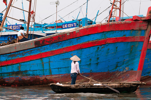 ranquil scene in Cat Ba Bay: A woman in a traditional Vietnamese conical hat rows past a vibrant blue and red boat