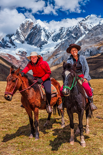 Young girl riding a horse with a young female tourist, Peruvian Andes, South America. Andes Mountain Range is located in South America, running north to south along the western coast of the continent.It is a continual range of highlands along the western coast of South America.The Andes extend from north to south through seven South American countries: Argentina, Bolivia, Chile, Colombia, Ecuador, Peru, and Venezuela, and is the longest continental mountain range in the world.
