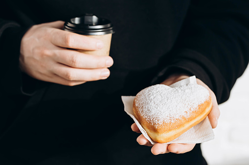 A paper cup of coffee and a heart-shaped donut in male hands on the background of a table in a cafe, close-up. Valentine's Day concept. Coffee break with donut.