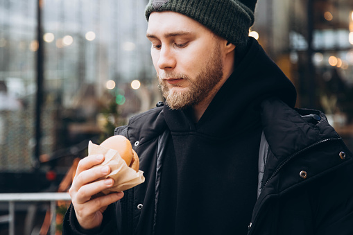 Attractive bearded young man wearing a jacket and hat with a burger outside. A man eats fast food in front of a cafe.