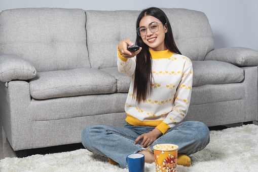 Beautiful Asian woman holding TV remote control Smiling happily while watching TV in the living room at home.