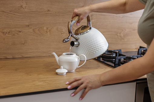 Close-up of a woman's hand pouring water from a textured white kettle into a ceramic teapot on a wooden kitchen counter.