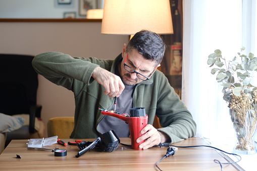 A man sitting at home on the dining table, by the window, wearing glasses, repairing a household appliance.