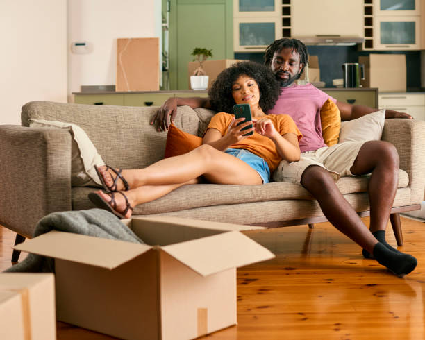 istock Couple Take A Break From Unpacking Boxes On Sofa Looking At Mobile Phone In New Home On Moving Day 2058262605