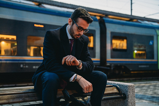 A young businessman is tired after a day at work. He's sitting on a bench at the station, waiting for the train.