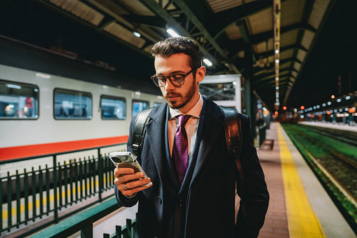 A young adult businessman is waiting for the train at the station, using his smart phone. He's wearing a full suit and a coat.