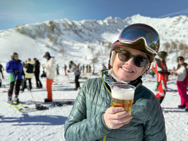 porträt einer lächelnden skifahrerin, die auf einer skipiste bier trinkt - beer ski apres ski snow stock-fotos und bilder