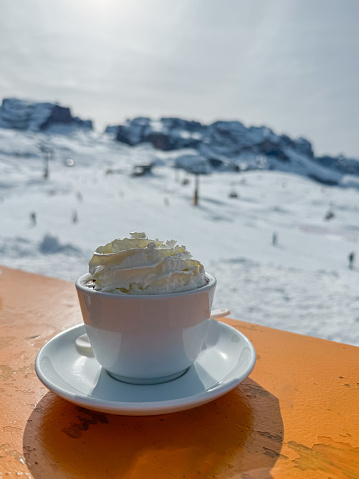 Close-up of coffee cup with whipped cream on wooden table at ski resort cafe. Coffee served on table at cafe with ski slopes in background.