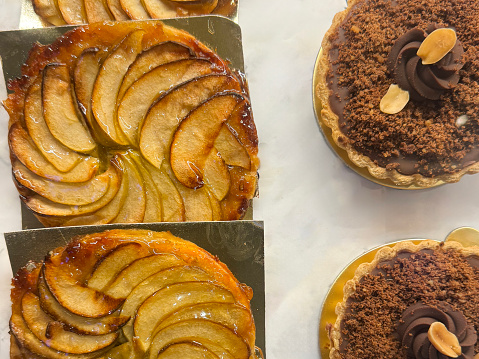 Stock photo showing close-up, elevated view of individual French patisserie apple tarte tatin made with cases of short crust pasty filled with sliced apple covered in caramelised sugar and chocolate tarts of golden pastry cases filled with chocolate ganache, on sale in a glass bakery display.