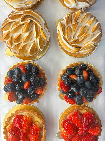 Stock photo showing close-up, elevated view of individual French patisserie fresh berry fruit and lemon meringue tarts made with cases of short crust pasty filled with creme patisserie custard, or lemon curd and topped with slices of strawberries and blueberries, or peaks of meringue, on sale in a glass bakery display.