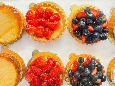 Stock photo showing close-up, elevated view of individual French patisserie fresh berry fruit tarts made with cases of short crust pasty filled with creme patisserie custard and topped with slices of strawberries and blueberries on sale in a glass bakery display.