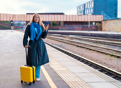 Woman in casual clothes with a yellow suitcase waiting for a train at the train station on the platform. Travel concept.