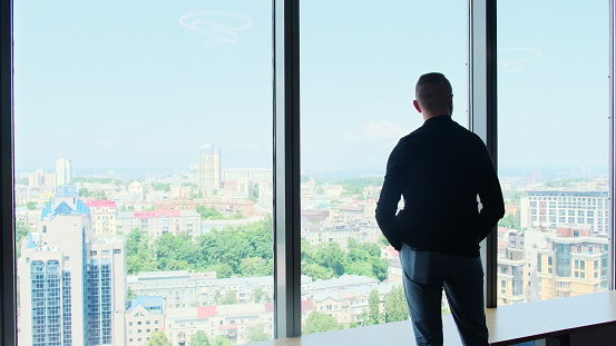 Brunette businessman stands near windows in company lounge zone. Male office worker stands next to panoramic window in company