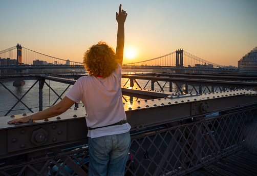 adult woman pointing upwards on the Brooklyn bridge at sunset, city skyline in the background.