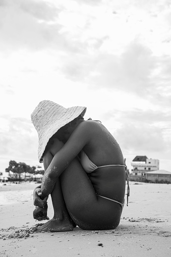 A young black woman in a bikini and a straw sun hat sitting on sand by the beach