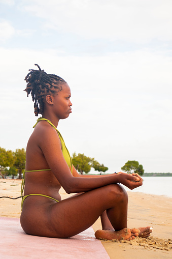A young black woman in a green bikini meditating on the beach
