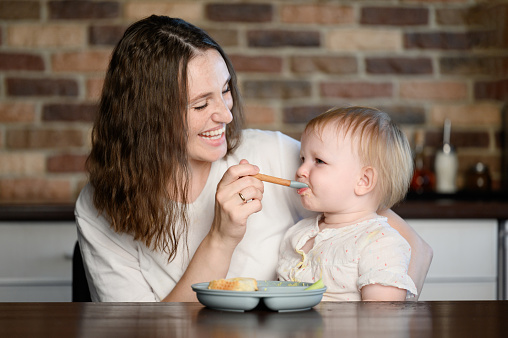 Happy mother feeding her kid puree mixture from a spoon in the kitchen. Healthy eating, baby weaning. Child tries his first solid food during lunch at home. Complementary feeding