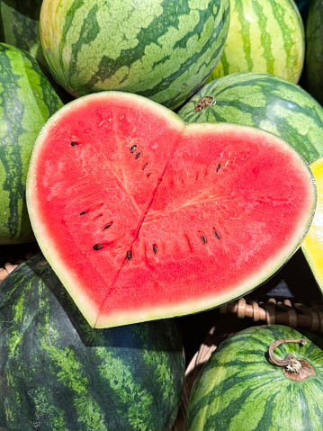 Stock photo showing close-up, elevated view of display of pile of fresh, whole watermelons with one sliced in heart shape revealing the bright red inner flesh and the rows of black pips / seeds.  Watermelons are particularly sweet and juicy, with more than 90 percent of the flesh being made up of water.