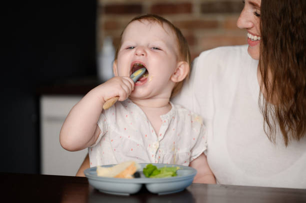 istock Charming child independently eats healthy chopped vegetable puree with a spoon while sitting in her mother arms. Self-feeding concept, complementary feeding 2058461774
