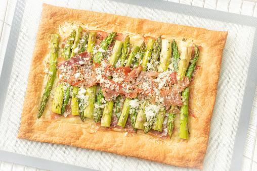 Homemade asparagus tart with puff pastry, emmental cheese, asparagus, olive oil, proscuitto,and garnishh with parmesan cheese, thyme leaves and baked in oven on a silicone baking mat