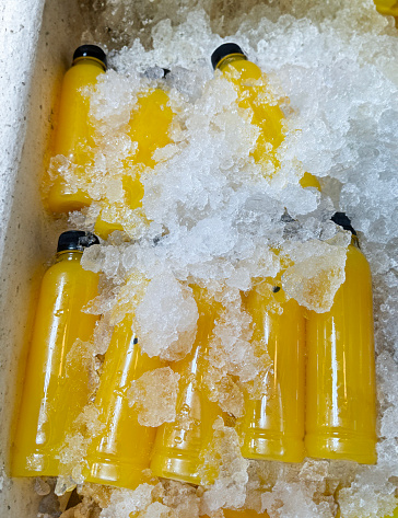 Stock photo showing close-up, elevated view of polystyrene box filled with ice cubes and containing plastic bottles of orange and pineapple juices being kept chilled.