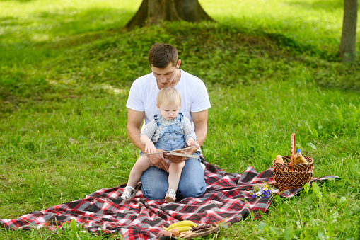 Young happy caring father reading a book to his child while sitting on a blanket during a picnic in a park.Spending time together,communicating,learning.