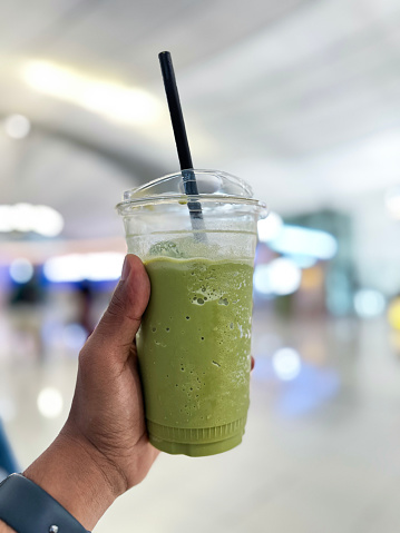 Stock photo showing close-up view of a lidded, plastic disposable cup with green drinking straw containing a green, matcha tea frappuccino drink held by unrecognisable person. This drink is made by blending ice, milk, vanilla syrup and green tea powder.