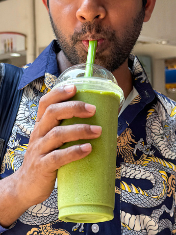 Stock photo showing close-up view of a lidded, plastic disposable cup with green drinking straw containing a green, matcha tea frappuccino drink held by unrecognisable person wearing dragon patterned short-sleeved shirt. This drink is made by blending ice, milk, vanilla syrup and green tea powder.