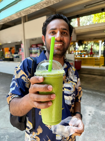 Stock photo showing close-up view of a lidded, plastic disposable cup with green drinking straw containing a green, matcha tea frappuccino drink held by Indian man wearing dragon patterned short-sleeved shirt. This drink is made by blending ice, milk, vanilla syrup and green tea powder.