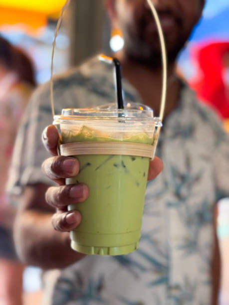 Stock photo showing close-up view of a lidded, plastic disposable cup with green drinking straw containing a green, matcha tea frappuccino drink held by unrecognisable person wearing palm leaf  patterned short-sleeved shirt. This drink is made by blending ice, milk, vanilla syrup and green tea powder.
