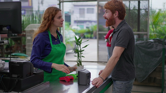 Male customer buying plant at horticulture cashier. Female employee wearing green apron interacting at checkout with client at local business store