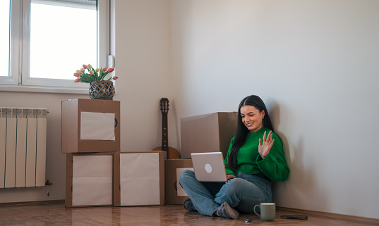 A young beautiful woman is sitting on the floor in her new apartment, using her laptop computer and talking on a video call. There are cardboard boxes around her.