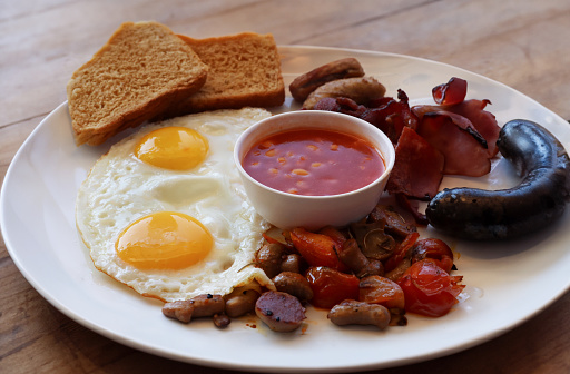 Stock photo showing close-up, elevated view of a white plate containing a fried breakfast with ramekin of baked beans, two sunny-side-up fried eggs, bacon rashers, sausages, grilled tomatoes, mushrooms, black pudding (blood sausage) and wholemeal toast.
