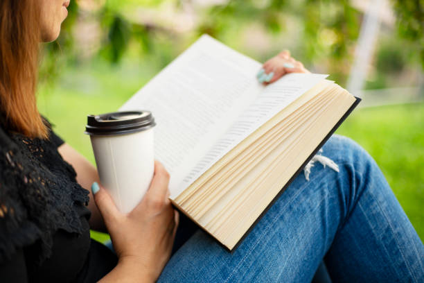 Woman reading book and holding coffee cup in park. Natural light candid shot with selective focus. Education and leisure concept. Design for poster, book cover, banner