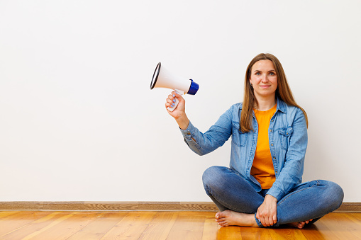 Casual young woman holding a megaphone while sitting cross-legged on a wooden floor, concept of communication.