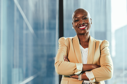 Confident black businesswoman in the office stands with her arms crossed, portraying success and professionalism. She exudes confidence and happiness as she smiles and looks at the camera.