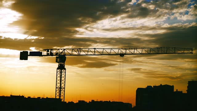 Silhouette of construction crane against beautiful yellow sunset. Mobile crane against evening cloudy sky. Construction works on industrial housing in city. Real estate, apartment block. New building.