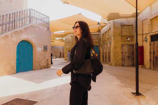 Handsome female in black casual clothes walking the old streets with authentic Persian buildings at Al Bastakya district of Dubai city, United Arab Emirates