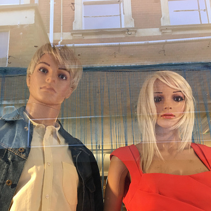A close-up of a shop window with a male mannequin positioned to the left at an angle, with short blonde hair modelling a blue denim jacket and plain cream or white coloured shirt with a collar. A female mannequin with blonde longer hair in a sleeveless bright red dress is to his right. In the Norwich charity shop window, you can see the reflection of the building opposite, the guttering and windows