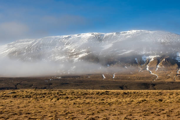 Yellow grass on a field. Mountain partly with a snow, partly visible the volcanic dark rock. Light fog hiding the mountain. Bright blue sky. Area of Hvitserkur, North-West Iceland.