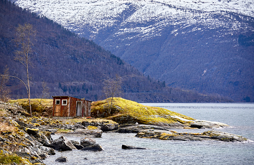 Small shed on an island in Norway, the is a bird in the sky an in the back you see the with snow covered mountains.