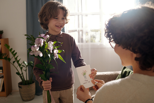 Happy cute boy with bunch of flowers passing handmade greeting card to his mother while congratulating her on traditional spring holiday