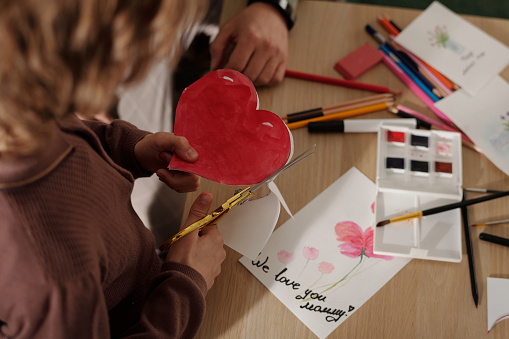 High angle of hands of child cutting out red paper heart over table with crayons, handmade postcards, watercolors and highlighters