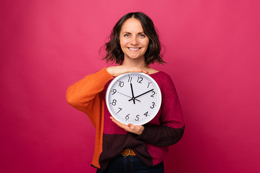 Studio portrait of a beautiful smiling middle age woman holding a round clock over pink background.