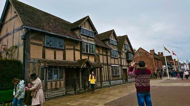 un hombre tomando una foto de una mujer frente a la casa natal de shakespeare bajo un cielo nublado con peatones que pasan - william shakespeare poet literature history fotografías e imágenes de stock