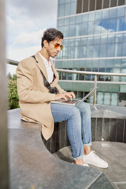 front view of middle eastern young man using laptop outdoors, working online or web surfing, copy space. indian guy sitting on bench on street, typing on computer keyboard - surfing wireless vertical outdoors lifestyles imagens e fotografias de stock