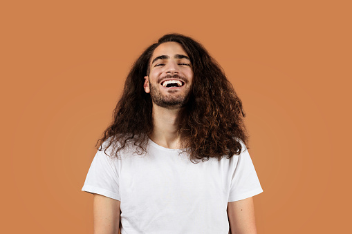 Joyful young Hispanic man with curly brown hair laughs and smiles in casual pose, standing against beige studio backdrop, wearing white t-shirt. Guy expressing positive emotions and happiness