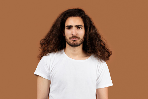 Studio Portrait Of Sad Arabic Guy With Long Curly Hair Standing On Brown Beige Background. Upset Young Man In Casual White T-Shirt Posing Looking At Camera Having Problems