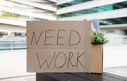 Cardboard sign with 'Need Work' text alongside personal items standing outdoors against modern business center on background, conceptual image for unemployment and joblessness problem